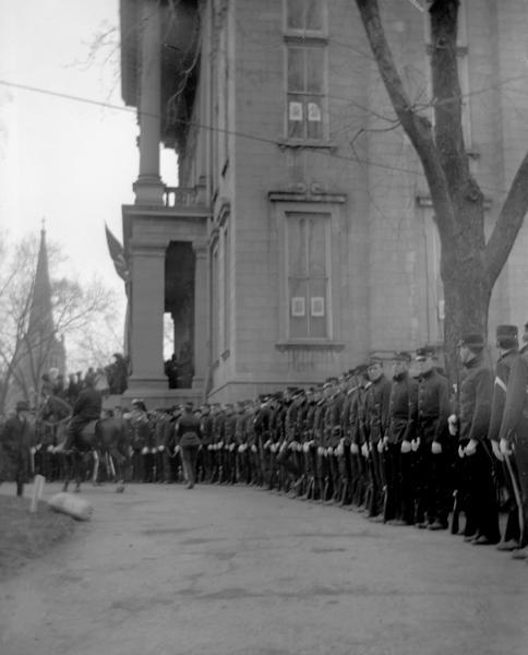 Military honor guard standing outdoors at the funeral of former Governor and Civil War hero Lucius Fairchild, which was held at the Wisconsin State Capitol on May 26, 1896. The funeral was reportedly the most magnificent ever staged in Wisconsin history. Fairchild's body lay in state in the Capitol, and special services for the public were held in the Capitol Park. Afterwards, Fairchild was buried at Forest Hill Cemetery.