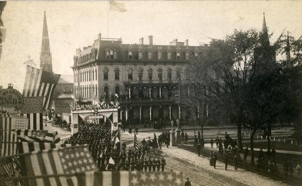 Military parade on Main Street honoring President Grover Cleveland's visit to Madison. Soldiers of the University Battalion are marching on West Main Street on their way to meet the President. The parade route is decorated with flags and a patriotic archway with a sign that reads, "We Greet You." In the background is the Park Hotel, on the right is the Capitol Park, and the steeple of St. Raphael's Catholic Church is behind trees.