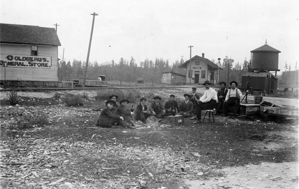A group of men near the Koss railroad depot, the majority of whom are seated on the ground. One of the men is sitting on a piano stool. To the right of the group is a hand-pump on a wooden platform. All of the men are drinking beer. A horse and wagon is standing near a general store on the left in the background. There is a large water tower on the right near the railroad tracks.