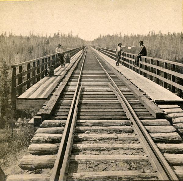 Stereograph of the view down the railroad tracks, with two men standing and one man sitting on the railing of the White River bridge, six miles south of Ashland. This bridge was part of the Wisconsin Central Railroad line, and was 1600 feet long and 110 feet above the water, making it one of the largest trestle bridges of its day.