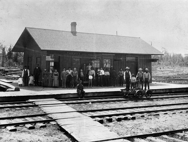 Elevated view across railroad tracks of the Wisconsin Central Railroad depot. Virtually the entire population of Sherry is thought to be standing on the platform for this photograph. A boy and three men are standing on a handcar in the foreground. At Sherry, passengers on the Wisconsin Central could make connections to the backwoods on logging trains.