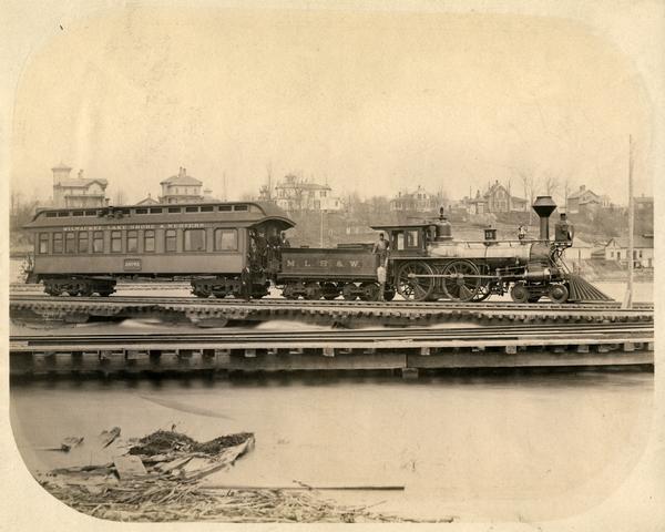 View across water of a Milwaukee, Lake Shore, and Western Railroad locomotive (No #33) pulling a pay car across a trestle over the Fox River. Several large homes are on a hill in the background.
