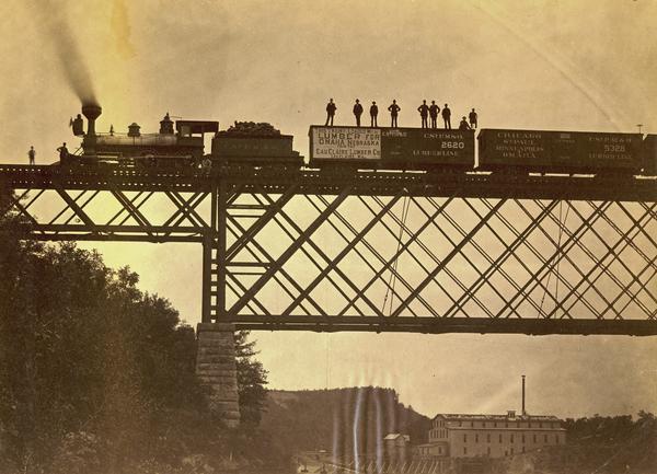 View looking up towards the special train of the Chicago, St. Paul, Minneapolis & Omaha Railroad comprised of 24 freight cars loaded with lumber and other wood products. A group of men are standing on top of one of the boxcars. The train was en route from Eau Claire to Omaha, Nebraska. In this photograph the train is crossing a bridge over the Chippewa River at Eau Claire. In the background is a large brick building, and a bluff.
