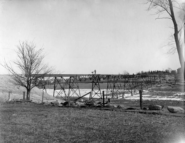 View across field towards the railroad bridge, seen with the construction almost completed. A man is standing in the middle of the bridge, and two or three other men stand just below him on beams. Another man wearing a long coat stands on the ground below. There is snow on the ground, and buildings are in the background on the far shoreline.