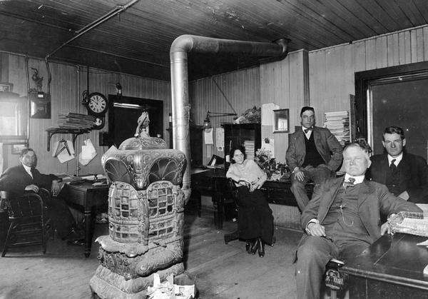 Employees of the Chicago, Milwaukee & St. Paul Railway in the company's Waukesha office.  Left to right they are Carl Mix, Ruth McGeen, Clarence Mica (seated on the table), Tom Taylor (seated in the chair), and John Lawless.  The room is heated by a large stove in the center of the room.