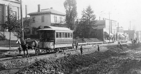 View across road construction towards a horse-drawn trolley on railroad tracks on Racine Street. Men and children are standing near piles of road building materials near the trolley.