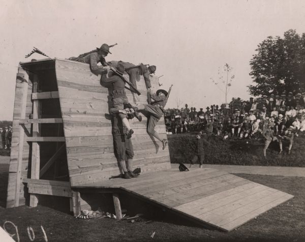 World War I recruits scramble over an obstacle during their training, while others watch in the background.