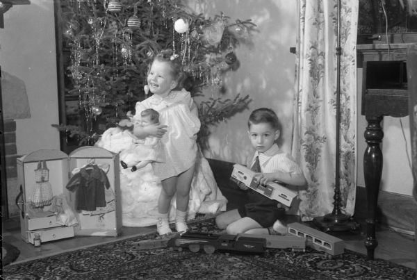 Roger and Mary Sue Mallory, in front of a Christmas Tree, playing with toys their father Lieut. Col. Robert Mallory, stationed in France, sent to them. Roger's train includes a wooden car with the famous World War I slogan: "40 hommes ou 8 chevaux." Mary Sue's doll "Mademoiselle" came from France with a trunk of clothes. A Christmas tree is in the background.