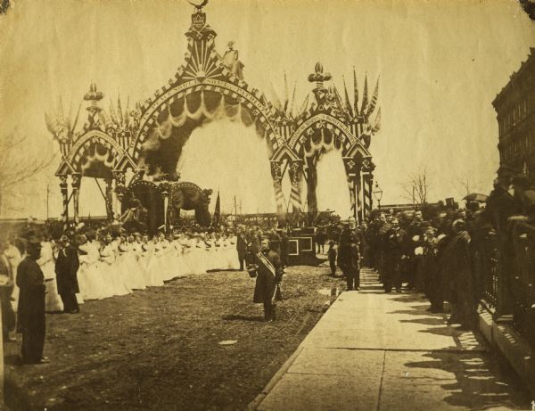 Albumen print of Abraham Lincoln's funeral procession. Thirty-six maidens in white surround Lincoln's hearse as it passes through the funeral arch, probably located at the intersection of Twelfth and Michigan Avenue, on its way into the city. Dignitaries and spectators watch the procession. Lincoln's funeral train is behind the arch.