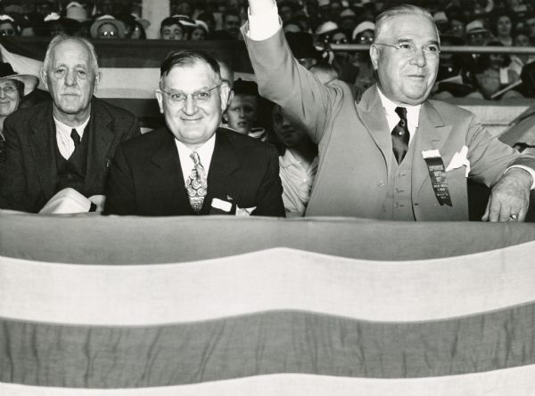 While watching the finish of a horse race at the Wisconsin State Fair, Governor Julius Heil raises his arm to cheer. Seated beside him (left to right) are Lieutenant Governor Walter S. Goodland and Col. Edward A. Kickhaefer.