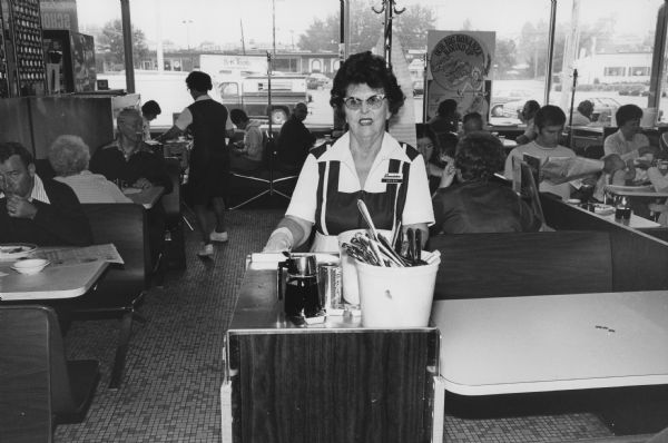 A waitress identified only as Helen clears dirty dishes from the tables at the Rennebohm Drug Store No. 19 restaruant, 204 Cottage Grove Road. This is the present site of the Madison Public Library's Pinney Branch. It is a busy morning and all of the other tables are full.