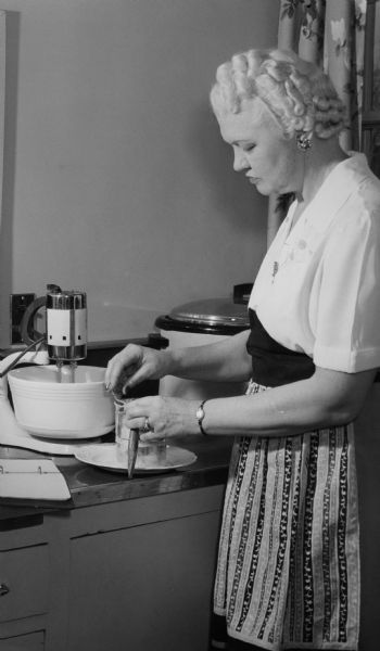 Mary Fowler Rennebohm wearing an apron and sifting flour.  Mrs. Rennebohm was the wife of Oscar Rennebohm, founder of the Rennebohm Drug Store chain and governor of Wisconsin.  It is not known if any of the Rennebohm drug store recipes can be attributed to Mrs. Rennebohm.  At one time the Rennebohm drug stores in Madison were so numerous that a favorite local joke identified the "Wisconsin" statue on the top of the Capitol as Mrs. Rennebohm "looking for a new site."