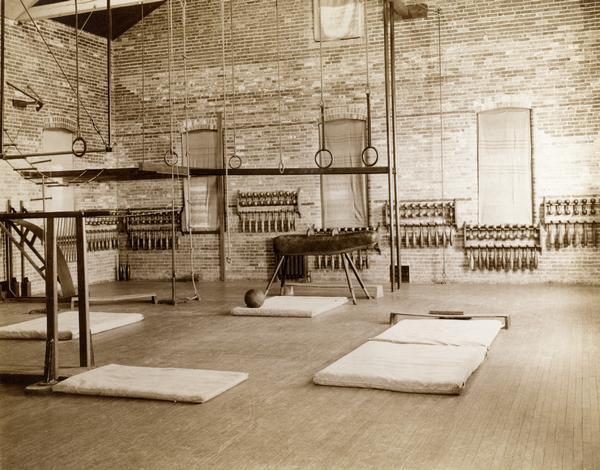 Gymnasium at the State School for the Deaf with floor mats, a pommel horse, rings apparatus, and racks of Indian clubs.