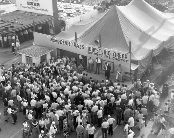 Elevated view of crowd of bystanders gathered in front of the tent of Jimmy Demetral's Wrestling Arena on the midway at the Wisconsin State Fair, as male wrestlers invite the audience to join in. A statement on the tent's sign challenges the bystanders, "We Meet All Comers."