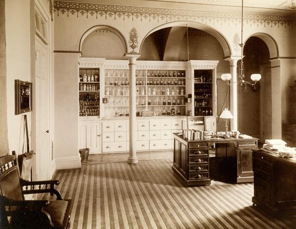 Pharmacy supplies are displayed in shelves in an office at the Wisconsin State Hospital for the Insane, lit by gas lamps hanging above the desk. (Mendota Mental Health Institute)