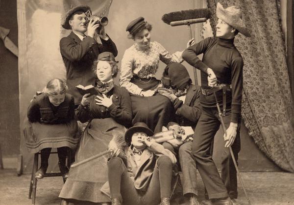 The Gesell family in costume with broom, sword, and other props for a humorous picture. Family members identified as follows: Gerhard Jr. (drinking from pitcher), Mrs. Gesell (seated with broom), Arnold (drawing sword-Civil War vintage), Gerhard Sr. (with stocking hat over head and mittens), Wilma (with book), Bertha (doubled over laughing), and Robert (sitting on floor with broom and bottle).