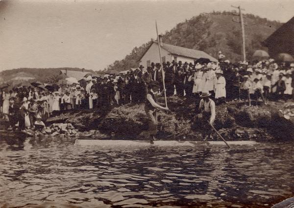 Log-rolling contest with large group of spectators on shore.