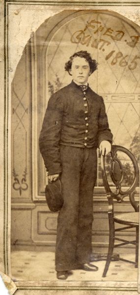 Full-length studio portrait of Fred D. Grant, son of Ulysses S. Grant, at 15 years of age, standing near a chair in front of a painted backdrop dressed in a military uniform. This image is one of several on a page from the Grant family bible.
