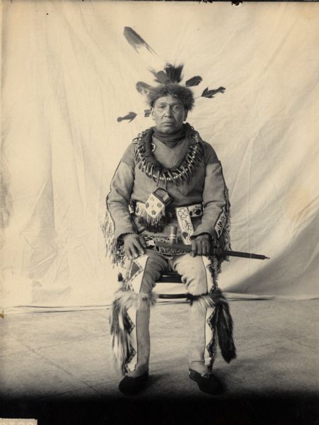 Portrait of Gegwejiwebinung or Gaygwachewaybinanung (Trying To Throw) or Gayshiqonnayyash (Swift Feather), Called Red Blanket, with Headdress and Bear Claw Necklace and Holding Pipe. Part of Algonquian and Chippewa (Leech Lake) Tribes
