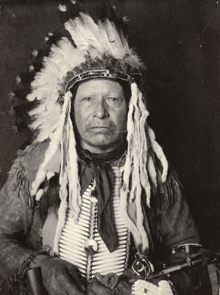 Studio portrait of Wakinyan Sapa (Black Thunder) in native dress with headdress and breastplate and holding a pipe and bag. Part of Siouan (Sioux) and Yankton Tribes.