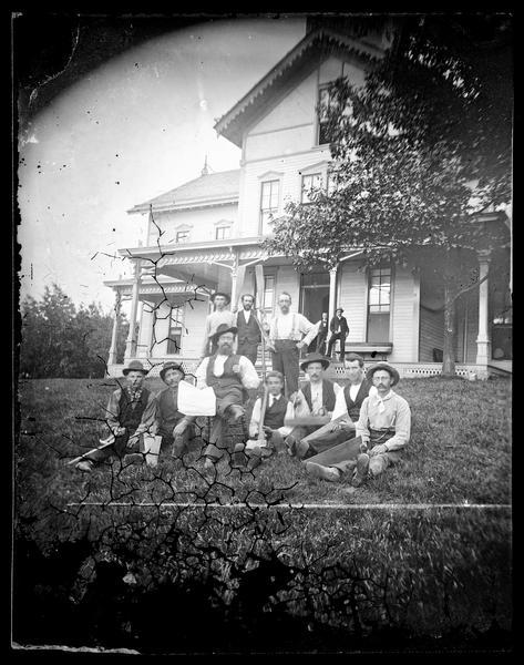 A group of men posing in the yard of the Tonyawatha House, later the Tonyawatha Spring Hotel, with paddles and carpentry tools. The hotel is a frame bracket-style structure that has latticework at the bottom of the porch and elongated windows overlooking Lake Monona. The hotel was under construction in 1879 and 1880.