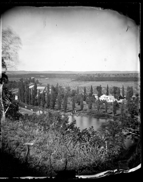 View from hill over a valley dotted with farmsteads and a meandering stream in the foreground. A group of three or four people are posing at the opposite shoreline. One or two people are sitting in a boat, another person is standing on the bank and holding an umbrella, and another person is standing nearby. Trees have been planted and carefully spaced along the shoreline, and a path through trees leads from the farmhouse on the right to farm buildings on the left. There is a road lined on both sides with a fence on the far left. Harvested fields are beyond, and hills are in the distance.