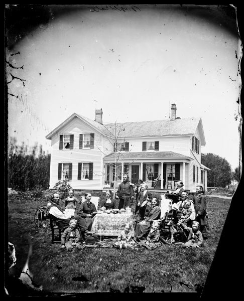 Norwegian family posing with their household possessions, in front of their home. The man dressed in military uniform is a Civil War veteran. Madison, Wisconsin (area), ca. 1870-1879. The family is in a yard around a table adorned with a lace cloth. Coffee cups, figurines and books are on the table. On the left is a man holding a newspaper that says "Norden." A baby sits in a buggy behind him. In the rear is a man dressed in a military uniform and, behind him, a man with a rake. On the right is a woman with a tray of beverages and, in front, a rocking horse.
