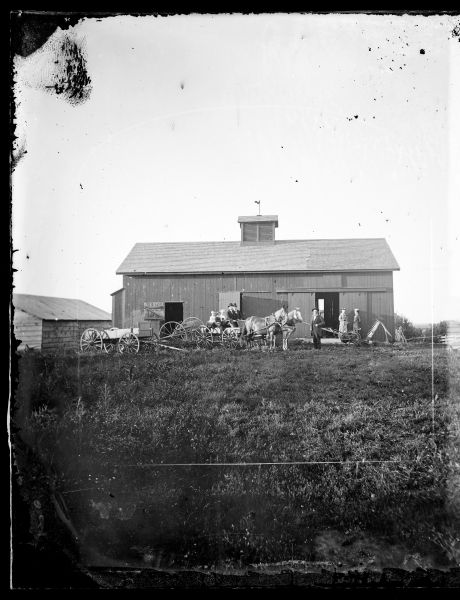 People are sitting in a horse-drawn wagon and standing next to farm machinery, including a dump rake and a hay mower.  A barn is behind them.