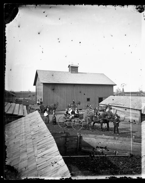 Family in Carriages with Barn Barnyard, with what appears to be a pigyard littered with corncobs. It is enclosed with a solid board fence and has slop troughs made from hollowed logs. Also in this farmyard view is a family in carriages and on horseback. A mower, dump rake, windmill and barn are in the background.
