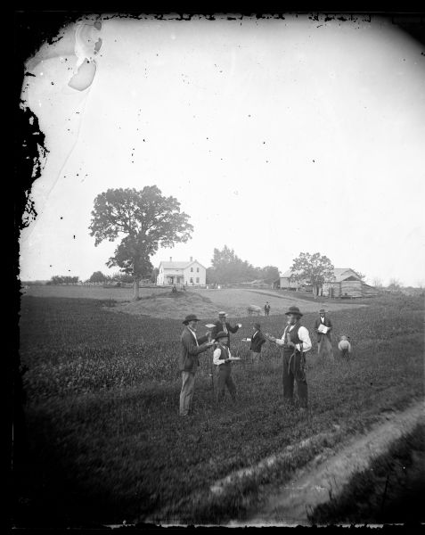 In this tableau, men and boys stand at the edge of the field in elaborate and fanciful poses. The man on the left holds a hoe and cake while the one on the right holds a harness, an ear of corn and a pretzel. In the distance, people sit on the haystack. A log animal shelter can be seen at the far right.