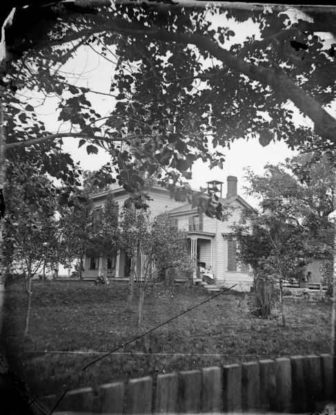 A man lies on the ground with a dog near the front door of his house. His wife and baby are posed on the steps of a side porch. The frame house has a stone foundation, shutters closed in a bay window on the side, a trellis in front, and a large dinner bell on the roof.