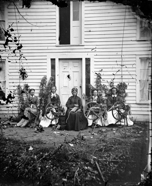Siri Rustebakke, center, sits in front of a house with her daughters and daughter-in-law and four spinning wheels. Original negative c109. Compare with D31-260. Siri Rustebakke and her husband, Amund, left the Begna Valley of Valdres, Norway, in 1866 and settled in Section 27 in the Town of Black Earth.  They moved to Webster County, Iowa, in 1875.