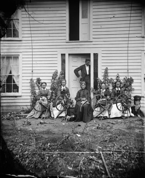 Amund Rustebakke, standing in doorway, and Siri Rustebakke, center, sitting, in front of a house with her daughters and daughter-in-law and four spinning wheels. A dog is sitting nearby on the right.