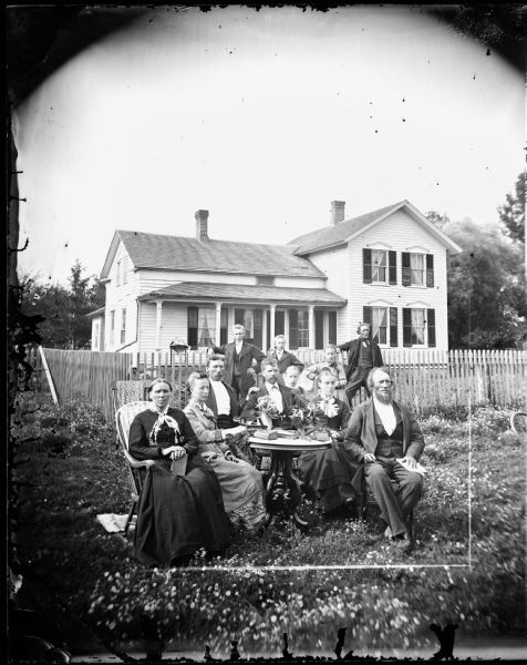 The Thor Brye family is posed in front of a fence in the yard. The women wear decorative scarves around their necks and one holds a stereoscope. The frame house behind them has shutters and cut work above its windows.