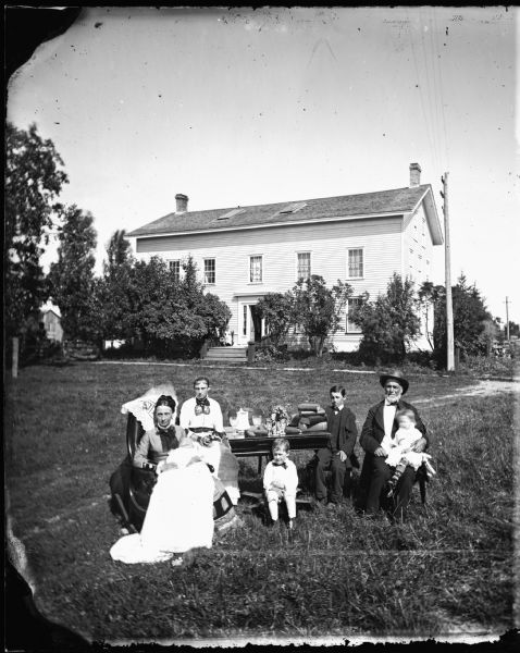 The William McFarland family is gathered around a table covered with books, pine cones, glassware and flowers in front of their Exchange Street house in McFarland, Wisconsin. Left to right are: Mrs. Sela Nelson McFarland holding Joseph; Martha, Edward, John; Mr. McFarland holding Mary, a step-daughter. A telegraph pole and wires are in the right background.