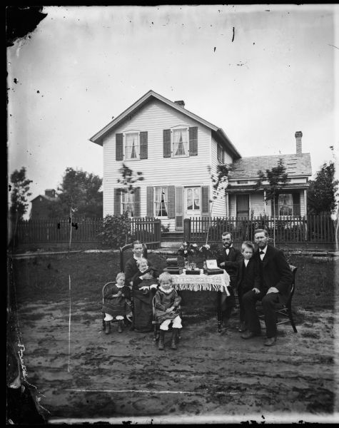 A family seated around a table that has a fringed cloth. The children sit in small chairs and a woman holds a baby. The frame house behind a picket fence has a "ten commandments" type window in its front door, and small windows on an addition. The front door has shutters along its side. Possibly the Anders Moldstad family.
