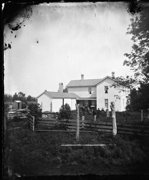 Stordock family within fenced yard playing croquet with frame house behind. There is a woodpile and small frame structure behind the house.