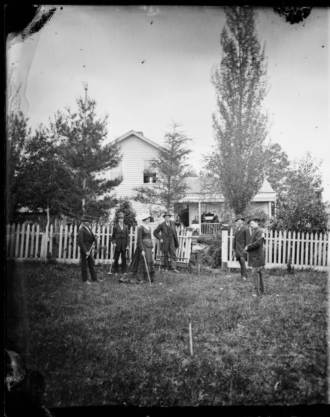 The home of R.D. Rickelesen. Five men and a woman in the foreground dressed in walking costumes are playing croquet with frame house behind; two women are on the porch near a parlor organ; there is a picket fence with the gate open behind croquet players.