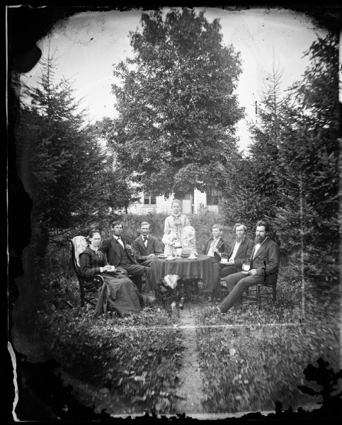The Reverend Peter Andreas Rasmussen (1829-1898) on the right, his wife Ragnhild Holland Rasmussen on the left and some visiting gentlemen, no doubt Lutheran ministers, are seated around a table drinking coffee. A large glass flower bell dome adorns the table. The frame house behind them is obscured by trees and tall grass.