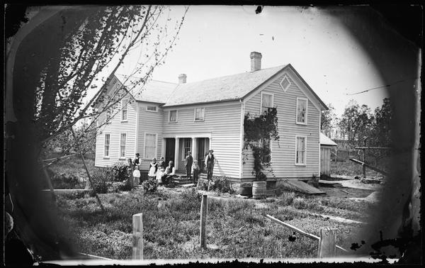 A family is standing and sitting in a yard of the porch of a upright and wing frame house that has a diamond shaped attic window, two doors in front and half windows above the porch. A water barrel, clothes washing kettle and a slanted basement entry door are on the right. A vine is growing near the corner of the house, partially covering two windows.