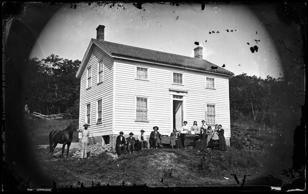 An extended family of fifteen stands in front of a wood frame house. On the left a man holds a black horse, and to the right of the doorway a woman sits at an organ with sheet music. A rough split-rail fence is in the background.
