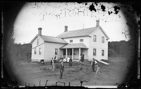 A family playing croquet in the yard of a frame house with three chimneys. A baby carriage is near the house on the left, and exterior cellar doors are on the right.