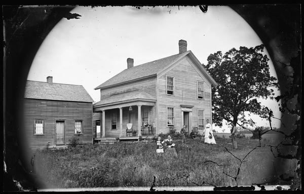 A family playing croquet in the yard of a side gable frame house with a porch. A birdcage is hanging from the porch and a young girl is clutching a doll. A rain barrel is near the door of an outbuilding on the right.