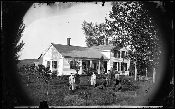 A family standing in a yard playing croquet, with a frame house and farm buildings behind them. An interesting rustic bench is on the right.