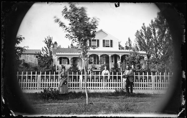 A man and two women are standing along a fence in the foreground, and another woman is standing on the porch. In the background is Samuel Barber's frame house with porch along front with carpenter's lace and trim at roof line. There are farm buildings in the background. Samuel Barber was the Middleton depot station master. A small dog is sitting on the fence between the two women.