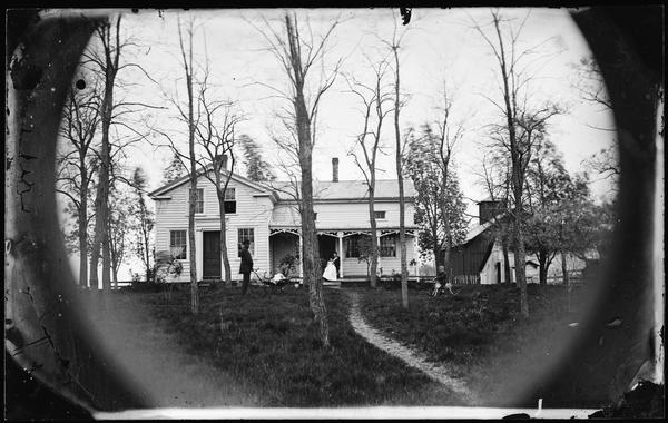 A man is standing on the far left with a baby in a baby buggy, a boy on the far right is sitting on a rocking horse on the lawn, and a woman holding baby is sitting on the porch of a frame Greek Revival house. The house has carpenter's lace on porch and two doors. Other frame farm buildings are in the background on the right. A winding path leads to the house through the grass and trees.