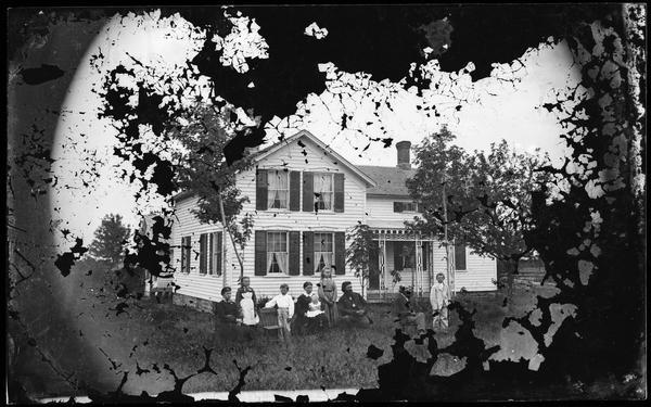 Family in yard, with a woman holding a baby. There is a girl with an apron-type dress with frame house behind that has nicely cut woodwork on porch. There is a stone foundation and small windows on second story in front and a small frame building behind house.