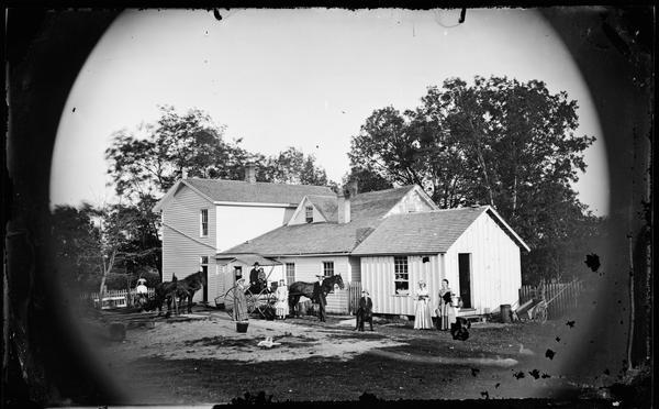 The Christian Keyser Preus family posing in the yard of the Spring Prairie parsonage with horse-drawn vehicles, and ducks. Behind them is a rural frame house with gable, and a man in a cart on the far left. Two women and a man are standing near a horse-drawn carriage in the center, in which a man is sitting with a child. On the right side is a boy, and two women, with one woman holding pails and a hat. C.K. Preus was the son of Herman Amberg Preus and was born in Spring Prairie in 1852. He was pastor at Spring Prairie from 1876-1897, and he died in 1921. 
