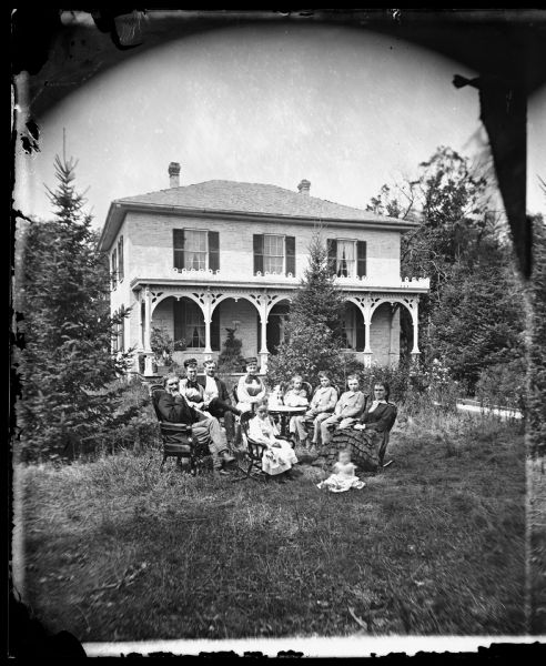 Family seated around table in yard with baby on grass and brick house with porch trim and shutters in background. This house is said to resemble the J. Hill house on Hubble Street in Black Earth, Wisconsin.
