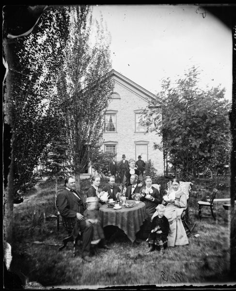 A group seated around a table in the yard drinking coffee. Pastor Even Johnson Homme (1843-1903), the founder of the Wittenberg Orphan's Home, is on the left. Mrs. Ingeborg Swenholt Homme sits at the right front. The photograph was made at the meeting of the Eastern District Norwegian Synod, held at the Immanuel Norwegian Evangelical Lutheran Church.